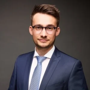 Porträt eines jungen Anwalts der Kanzlei mit Brille, blauem Anzug und Krawatte.